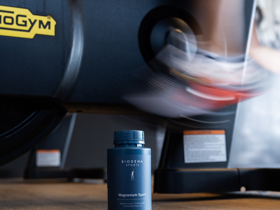 BIOGENA Magnesium Sport Supplement stands on the wooden floor in the background; someone is riding a stationary bike.