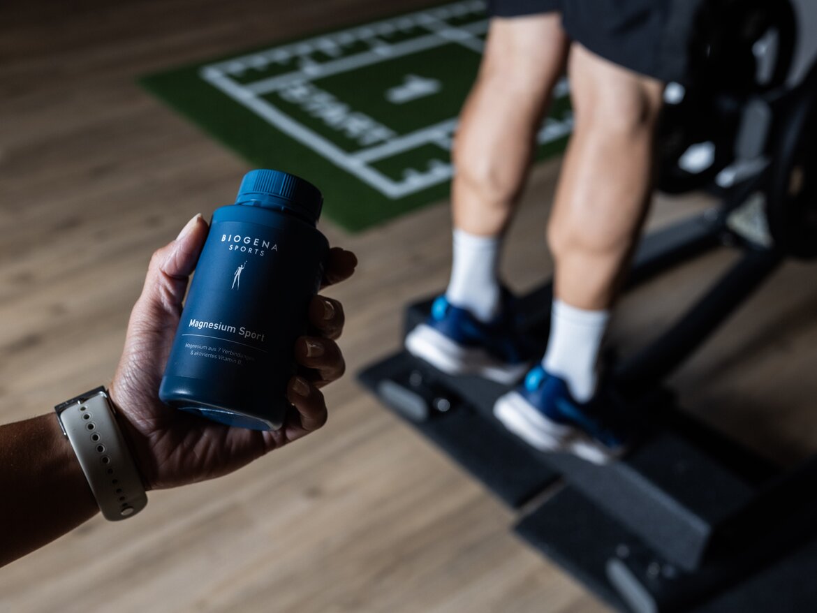 Biogena Magnesium Sport con B6 viene tenuto in mano mentre sullo sfondo qualcuno allena i polpacci.
