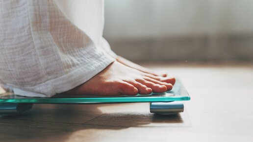 A person stands barefoot on a digital scale. Only the person's feet and part of the white fabric of their pants leg are visible. The floor is wooden, and the lighting in the room is soft.