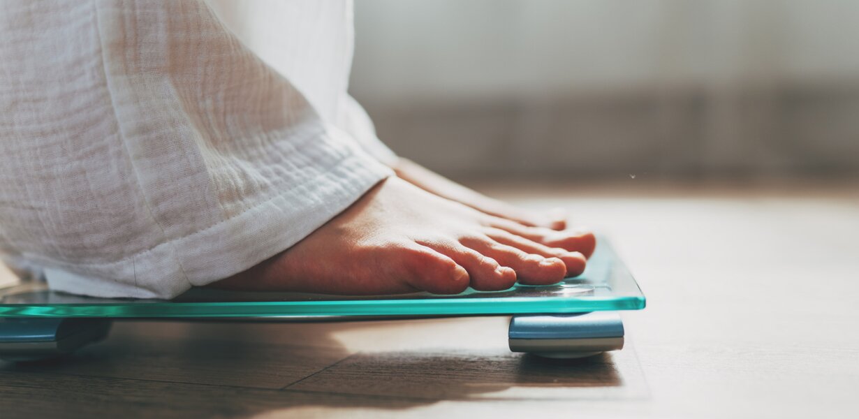 A person stands barefoot on a digital scale. Only the person's feet and part of the white fabric of their pants leg are visible. The floor is wooden, and the lighting in the room is soft.