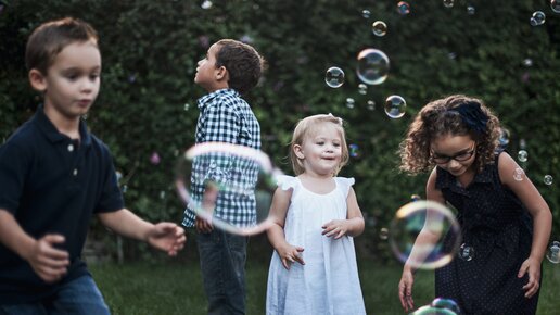 Four children are playing with soap bubbles in the garden. Two boys are looking up, while a girl in a white dress smiles and catches a bubble. The background is green and blurry.