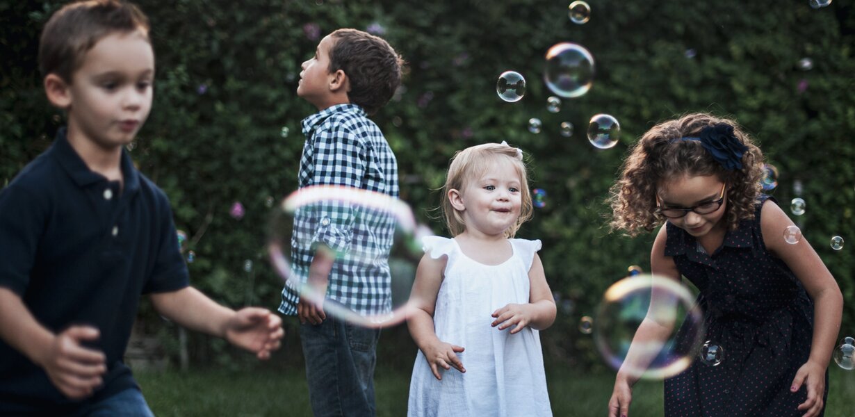 Vier Kinder spielen im Garten mit Seifenblasen. Zwei Jungen schauen in die Luft, während ein Mädchen in einem weißen Kleid lächelt und eine Blase fängt. Ein weiteres Mädchen mit lockigem Haar bückt sich, um eine Blase zu fangen. Der Hintergrund ist unscharf und grün.