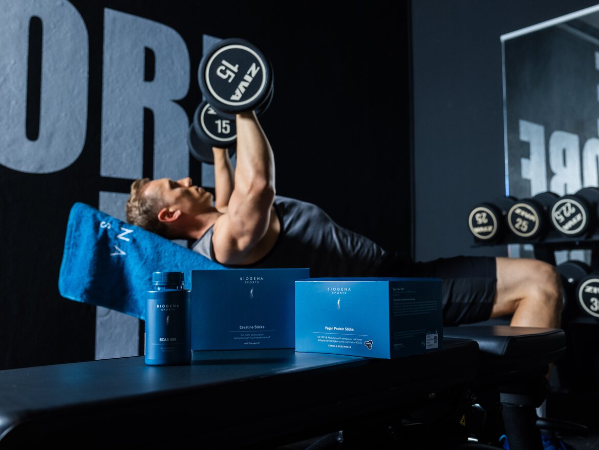 A man is training his upper body with dumbbells; in front of him stands the Biogena Muscle Bundle.