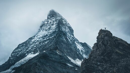 Berglandschaft mit dem verschneiten Matterhorn und einer Person, die auf einem Felsen am rechten Bildrand mit ausgebreiteten Armen steht.
