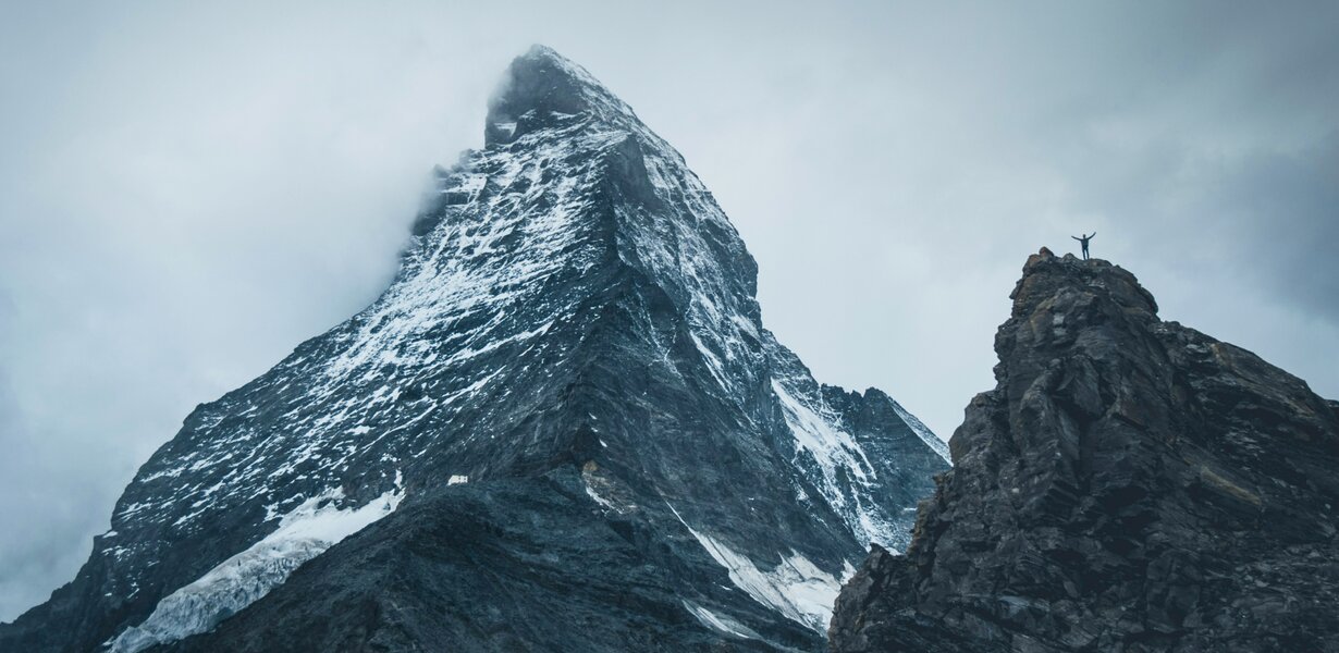 Berglandschaft mit dem verschneiten Matterhorn und einer Person, die auf einem Felsen am rechten Bildrand mit ausgebreiteten Armen steht.