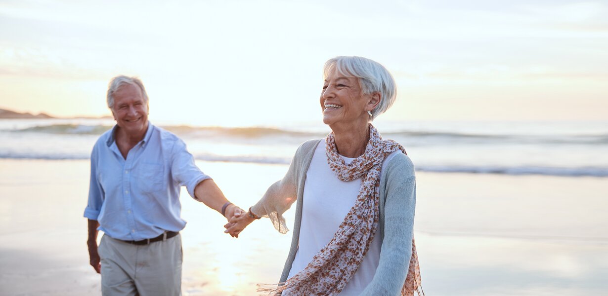 Älteres Paar lächelt und hält Händchen, während es bei Sonnenuntergang an einem sonnigen Strand entlanggeht.
