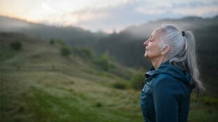 Ältere Frau mit grauem Pferdeschwanz, geschlossenen Augen, atmet ruhig in einer nebligen Hügellandschaft bei Sonnenaufgang.