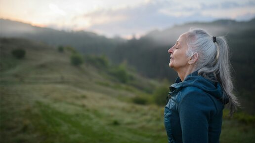 Ältere Frau mit grauem Pferdeschwanz, geschlossenen Augen, atmet ruhig in einer nebligen Hügellandschaft bei Sonnenaufgang.