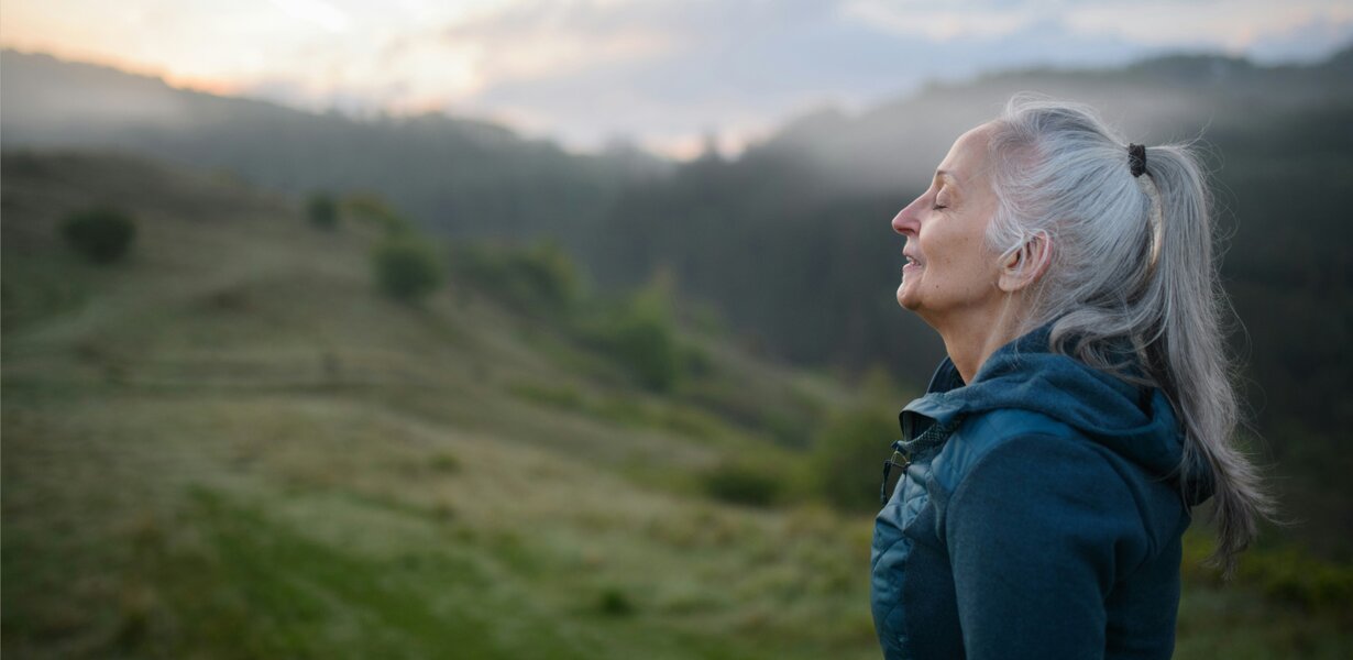 Ältere Frau mit grauem Pferdeschwanz, geschlossenen Augen, atmet ruhig in einer nebligen Hügellandschaft bei Sonnenaufgang.