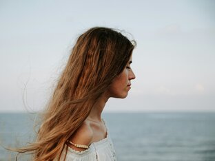 Frau mit langen, welligen Haaren, die ihre Augen schließt und vor einem Meer steht