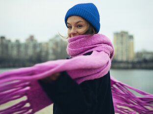 Frau mit blauer Mütze und pinkem Schal vor einem Fluss in der Stadt