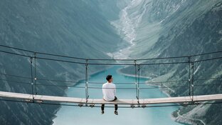 Mann der auf hoher Hängebrücke sitzt, mit türkisem See und Bergen im Hintergrund