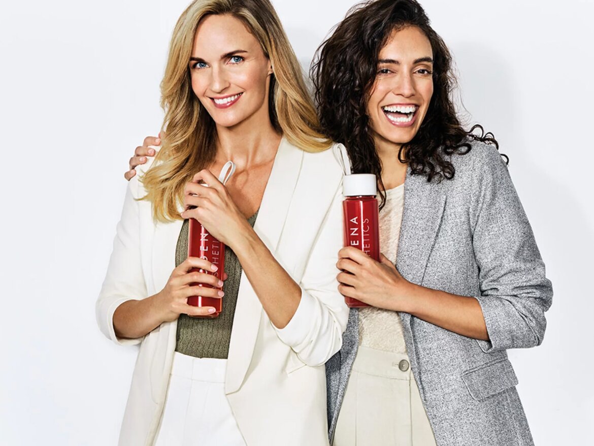 Zwei Frauen lächeln und halten jeweils eine Flasche BIOGENA AESTHETICS in den Händen. Beide tragen elegante Blazer in hellen Farben.