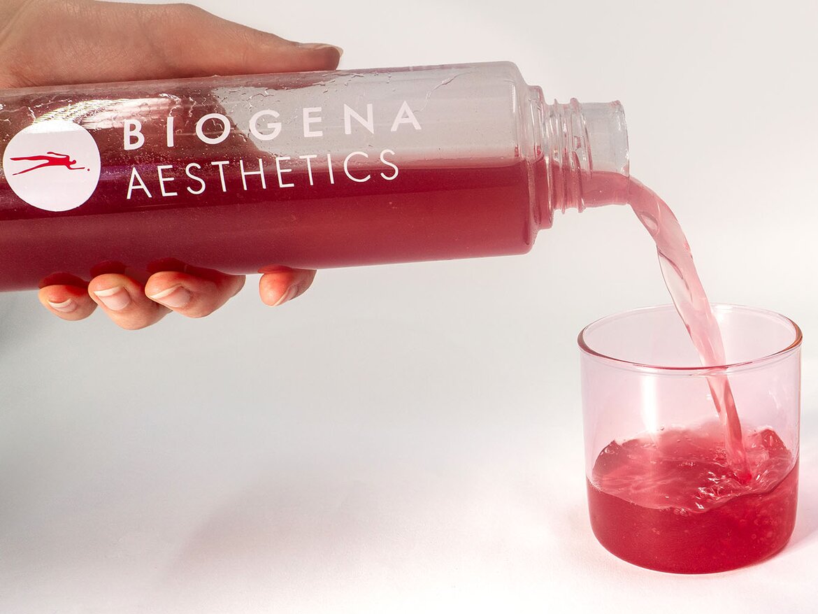 Una mano vierte una bebida roja desde una botella “BIOGENA AESTHETICS” en un vaso transparente.