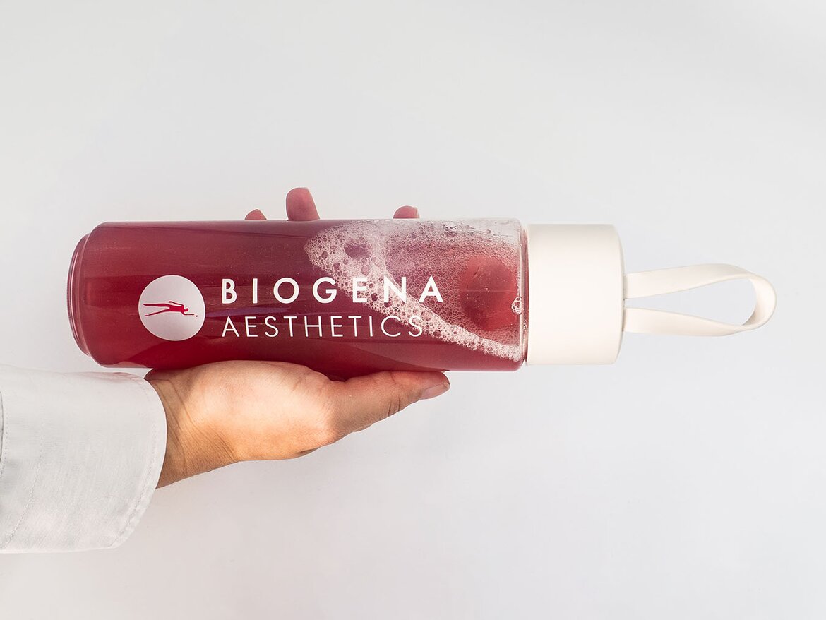 Una mano sostiene una botella transparente con líquido rojo y espuma, etiquetada “BIOGENA AESTHETICS”, tapa clara con asa.