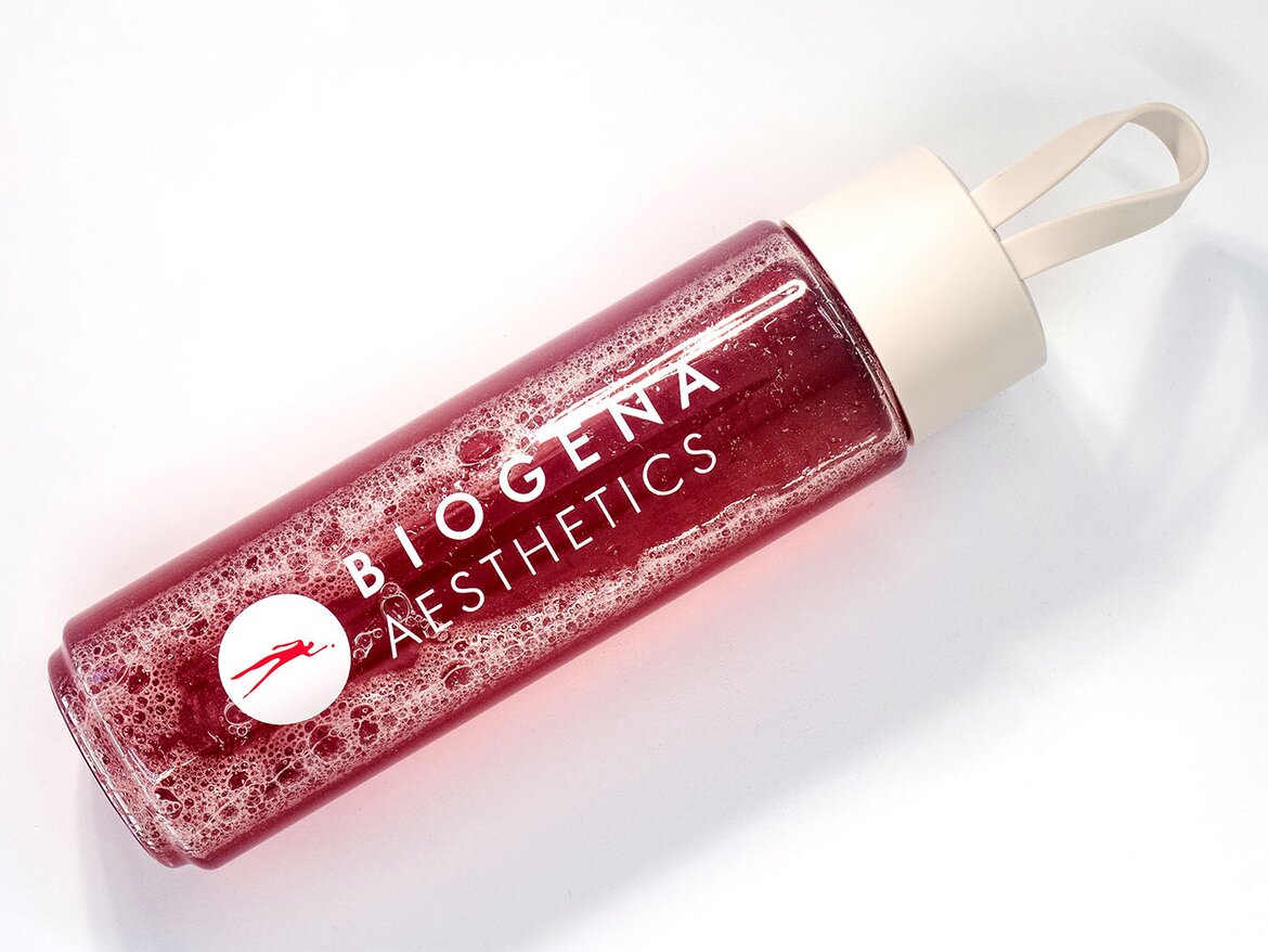 Botella transparente con líquido rojo y espuma, rotulada “BIOGENA AESTHETICS”, tapa plana clara con asa.