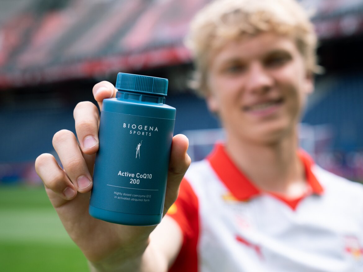 Młody mężczyzna w koszulce sportowej trzyma w dłoni opakowanie BIOGENA Active CoQ10 200 skierowane w stronę aparatu. W tle widać stadion piłkarski.