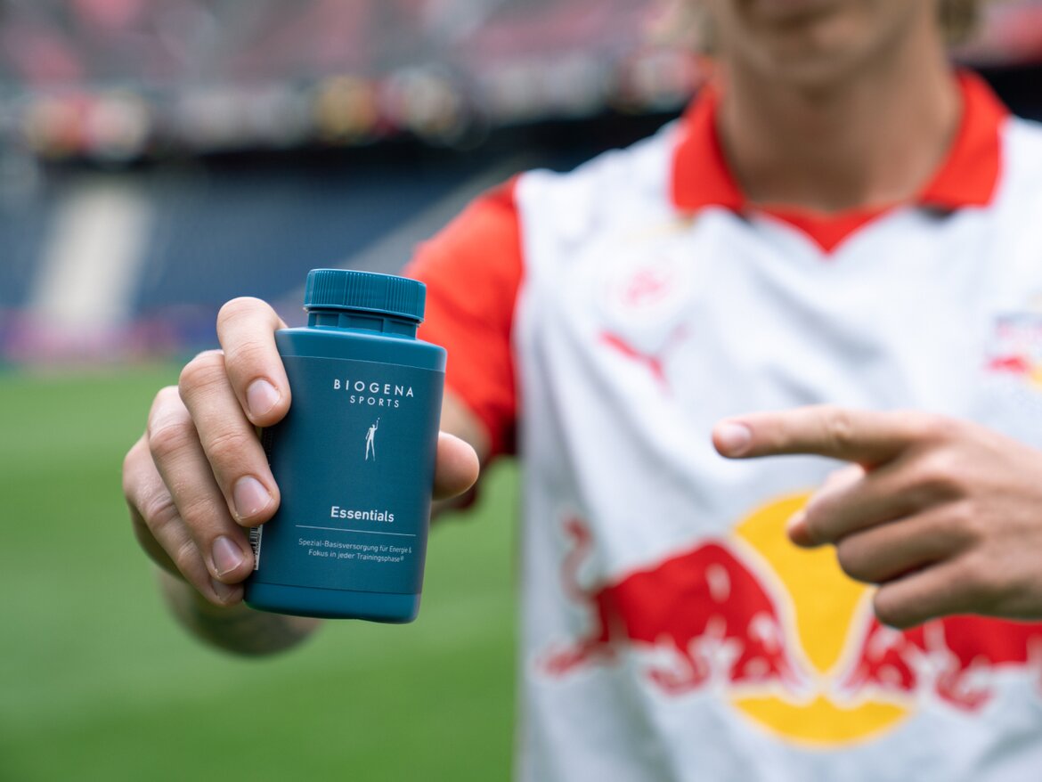 Tânăr îmbrăcat în tricoul unei echipe de fotbal ține în mână un flacon de BIOGENA Sports Essentials spre cameră și îl arată cu degetul. În fundal se vede un stadion de fotbal.