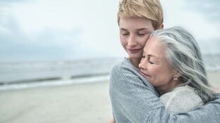 Eine ältere Frau mit grauen Haaren und eine jüngere Frau mit kurzen Haaren, die sich am Strand umarmen